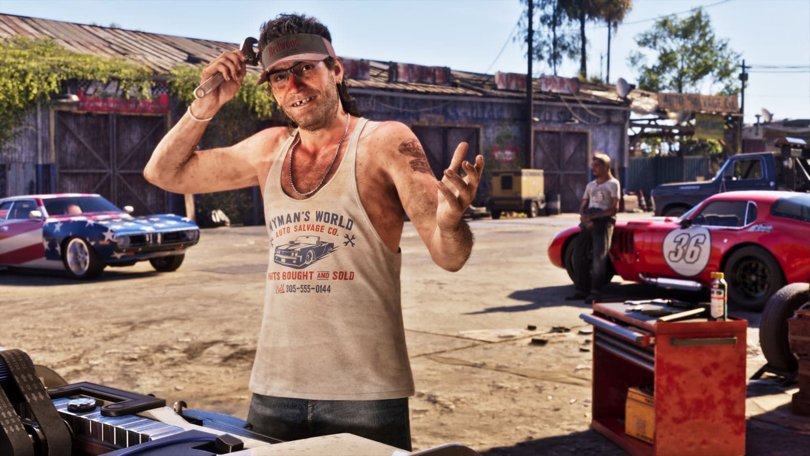 Wyman, in a Wyman’s World Auto Salvage tank top, holding a wrench in his salvage yard among classic cars